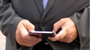 Un ejecutivo está consultando el móvil juntoa un portátil que tiene información financiera, pero la imagen solo muiestra el torso del ejecutivo, no su cara