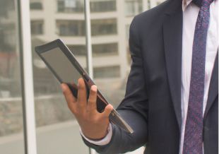 Un ejecutivo de empresa  revisa una tablet en la mano mientras se acerca al cristal de su oficina