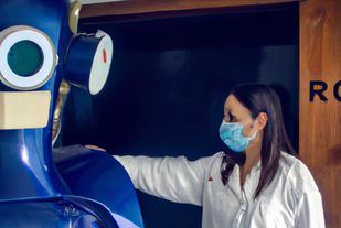 Una foto con un  robot, se da la mano con una enfermera, que lleva una mascarilla que le cubre el rostro en la puerta de un quirófano, 